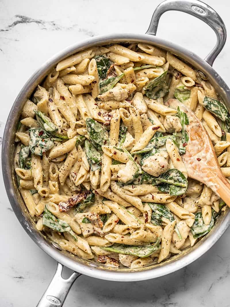 This super lush and Creamy Pesto Chicken Pasta is perfect for busy weeknights. Everything cooks in one pot and is done in under 30 minutes! BudgetBytes.com Overhead view of a skillet full of one pot creamy pesto chicken pasta with a wooden spoon in the side.